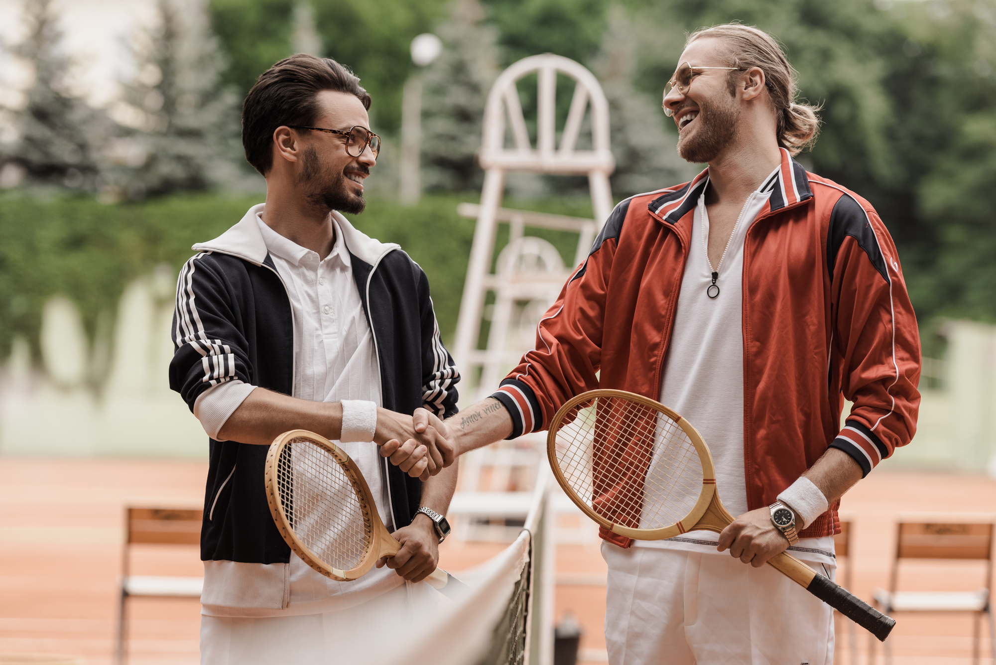 Two men, dressed in vintage athletic attire, stand on a clay tennis court shaking hands and laughing. One wears a black and white jacket, the other in a red jacket. Both hold wooden tennis rackets. The background features a chair umpire stand and greenery.