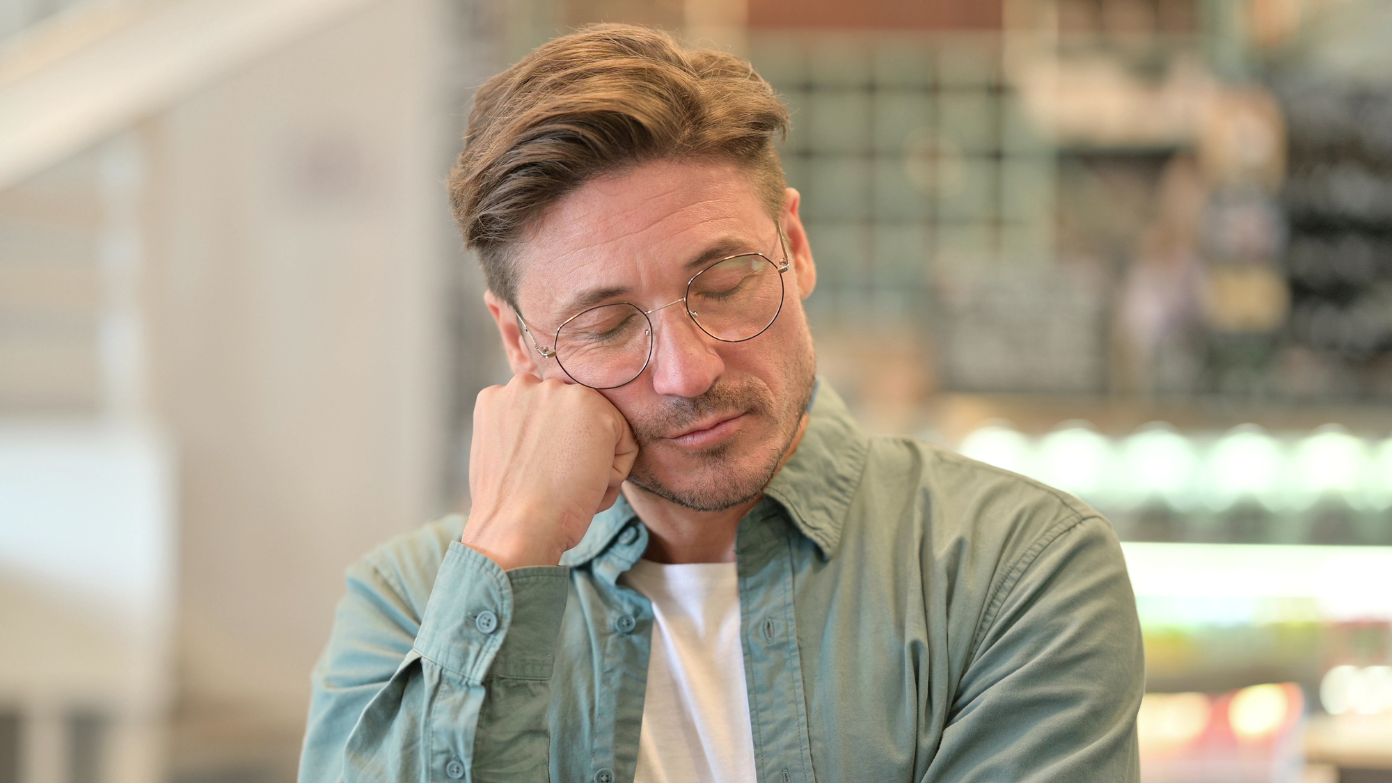 A man with light brown hair and glasses, wearing a green shirt over a white t-shirt, rests his head on his hand with his eyes closed. The background is blurred, showing an indoor setting with shelves and a possible menu board.