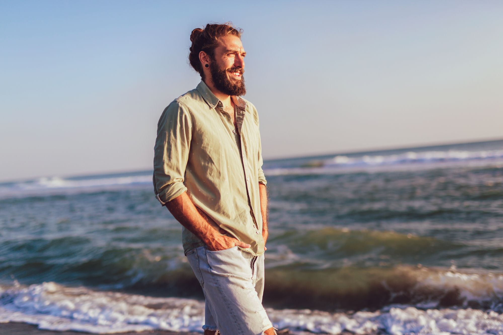 A bearded person with a man bun, wearing a light green shirt and rolled-up jeans, smiles while walking along the beach during sunset. They have their hands in their pockets and the ocean waves are visible in the background.