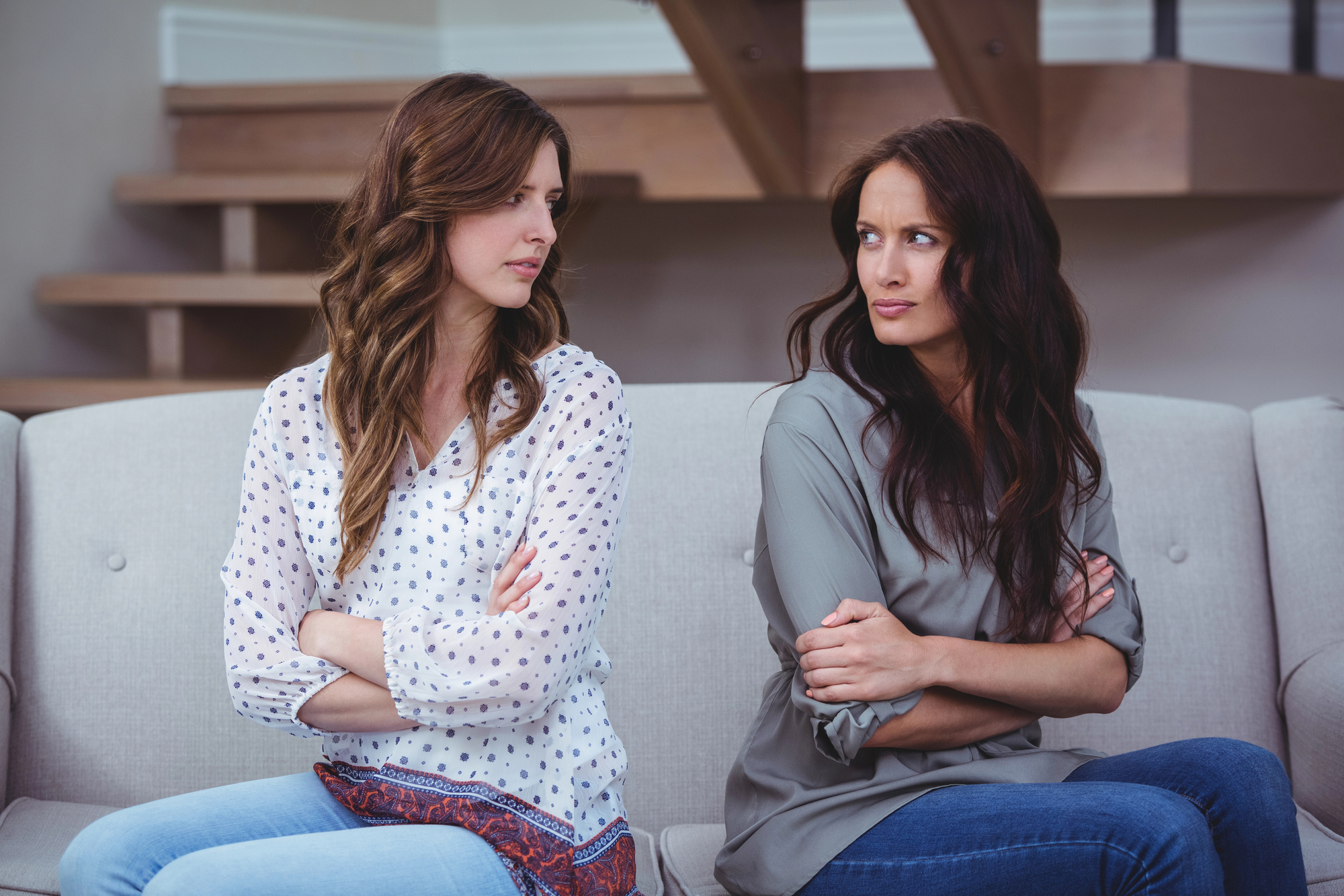 Two women sit on a couch facing each other with their arms crossed and serious expressions. One is wearing a white, patterned blouse, and the other a grey top. A staircase is in the background. Both appear to be upset or in a disagreement.