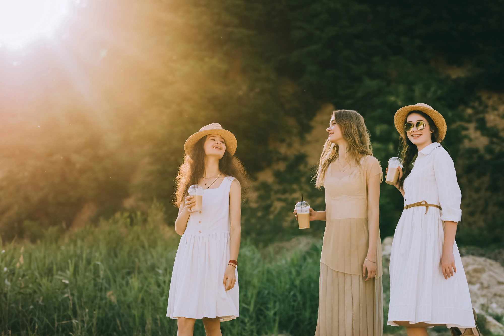 Three women stand outdoors in front of a forest, smiling and enjoying drinks in plastic cups. They are wearing summer dresses and wide-brimmed hats. Sunlight filters through the trees, creating a warm and bright atmosphere.