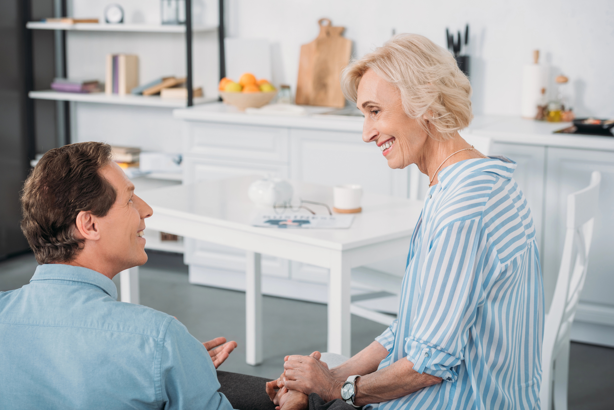 A smiling elderly woman with short blonde hair and a younger man with short brown hair are having a friendly conversation in a bright kitchen. The woman is wearing a striped blue shirt, and the man is in a light blue shirt. Kitchen items are visible in the background.