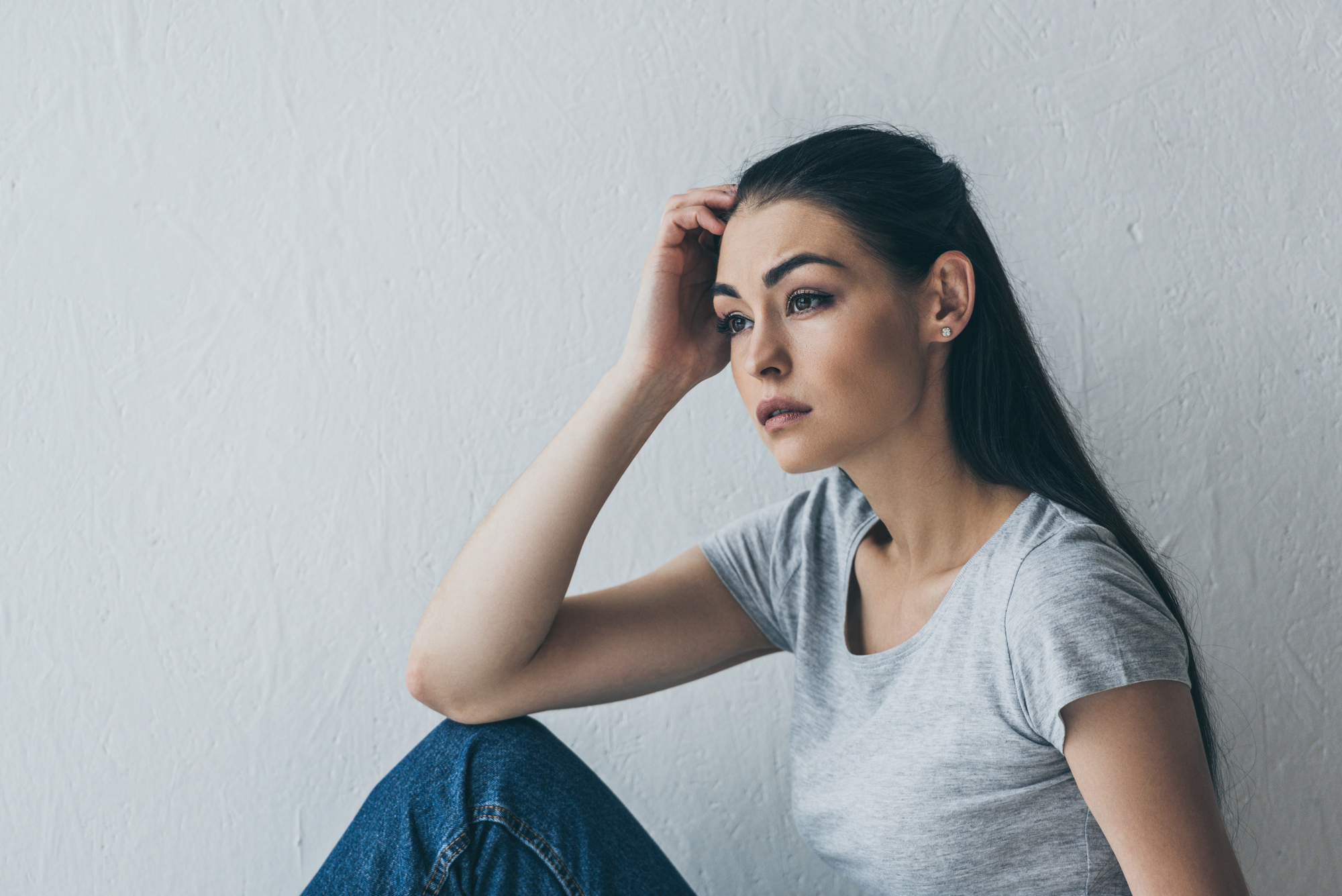 A woman with long dark hair in a gray t-shirt and blue jeans sits against a white textured wall. She looks pensive, with her head resting on her hand and her eyes gazing off into the distance.