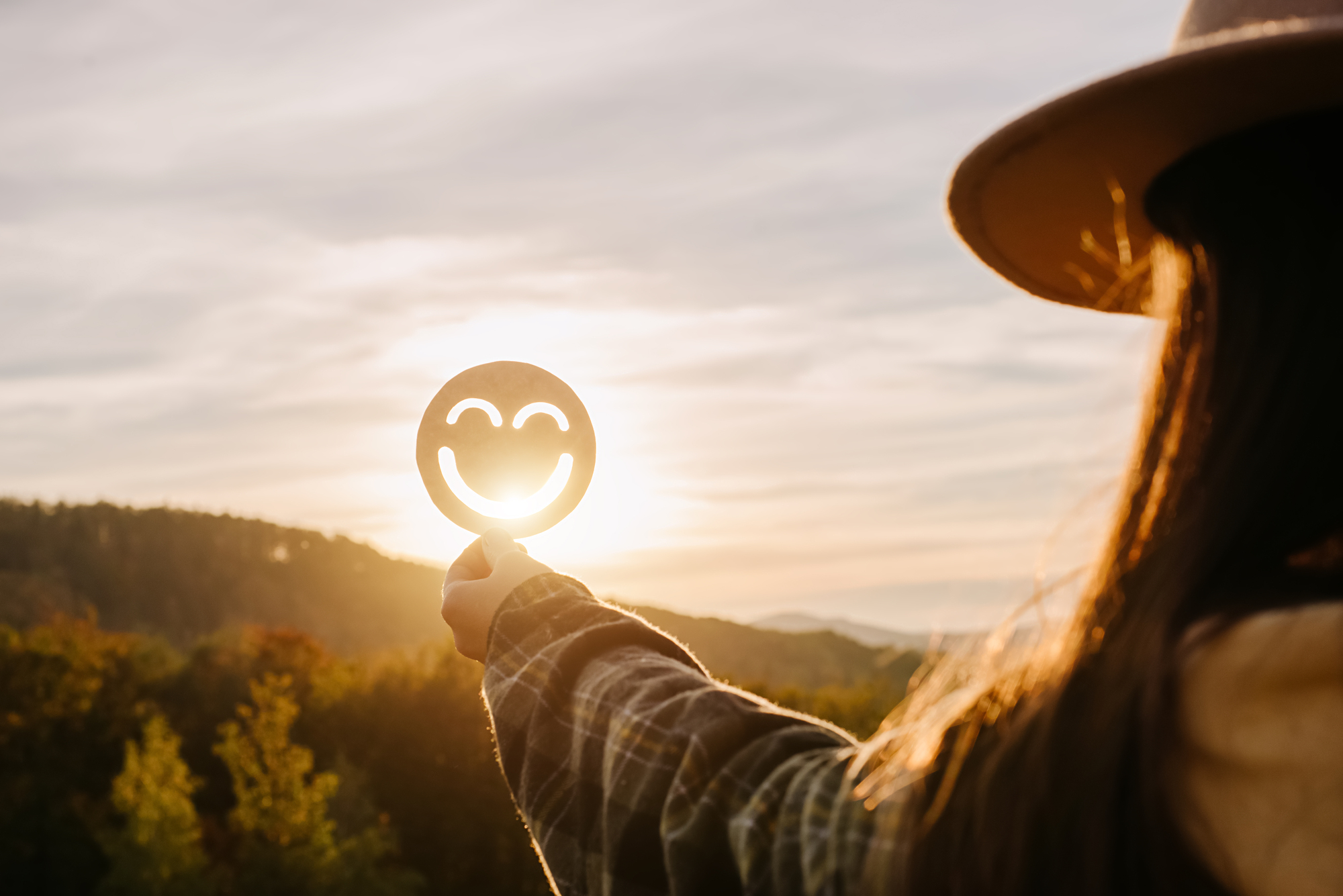A person holds up a circular cutout with a smiling face, positioned to catch the sunlight, creating a glowing effect. The person is wearing a hat and flannel shirt, standing in a scenic outdoor area with trees and rolling hills in the background.