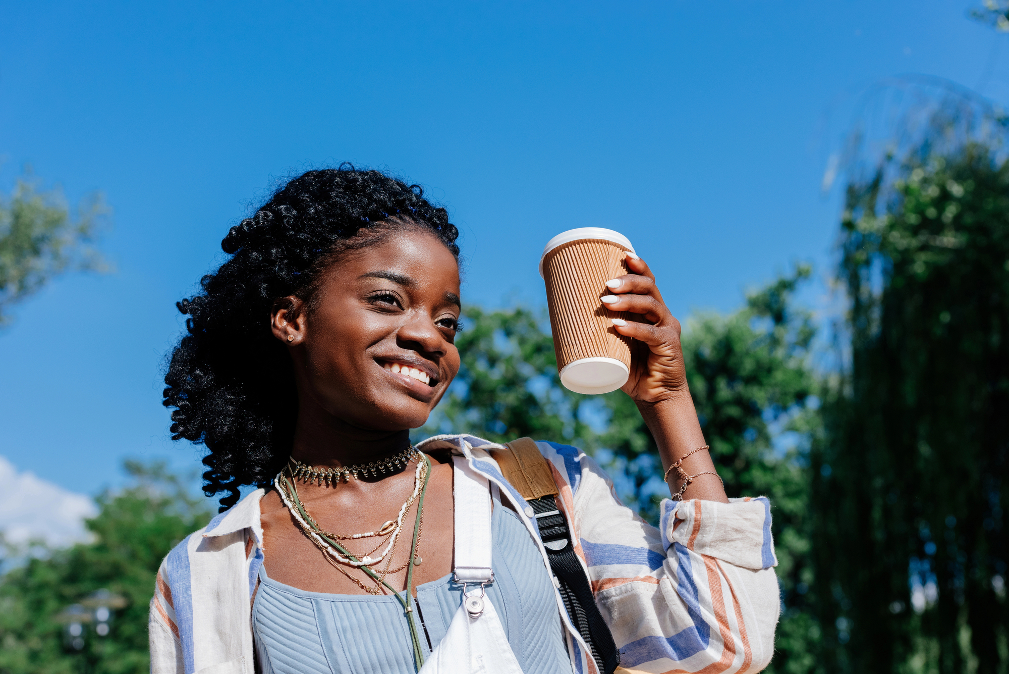A smiling woman stands outdoors against a clear blue sky, holding up a paper coffee cup. She is wearing a striped shirt over a blue top, layered necklaces, and a brown backpack. Green trees are in the background, indicating a sunny day.