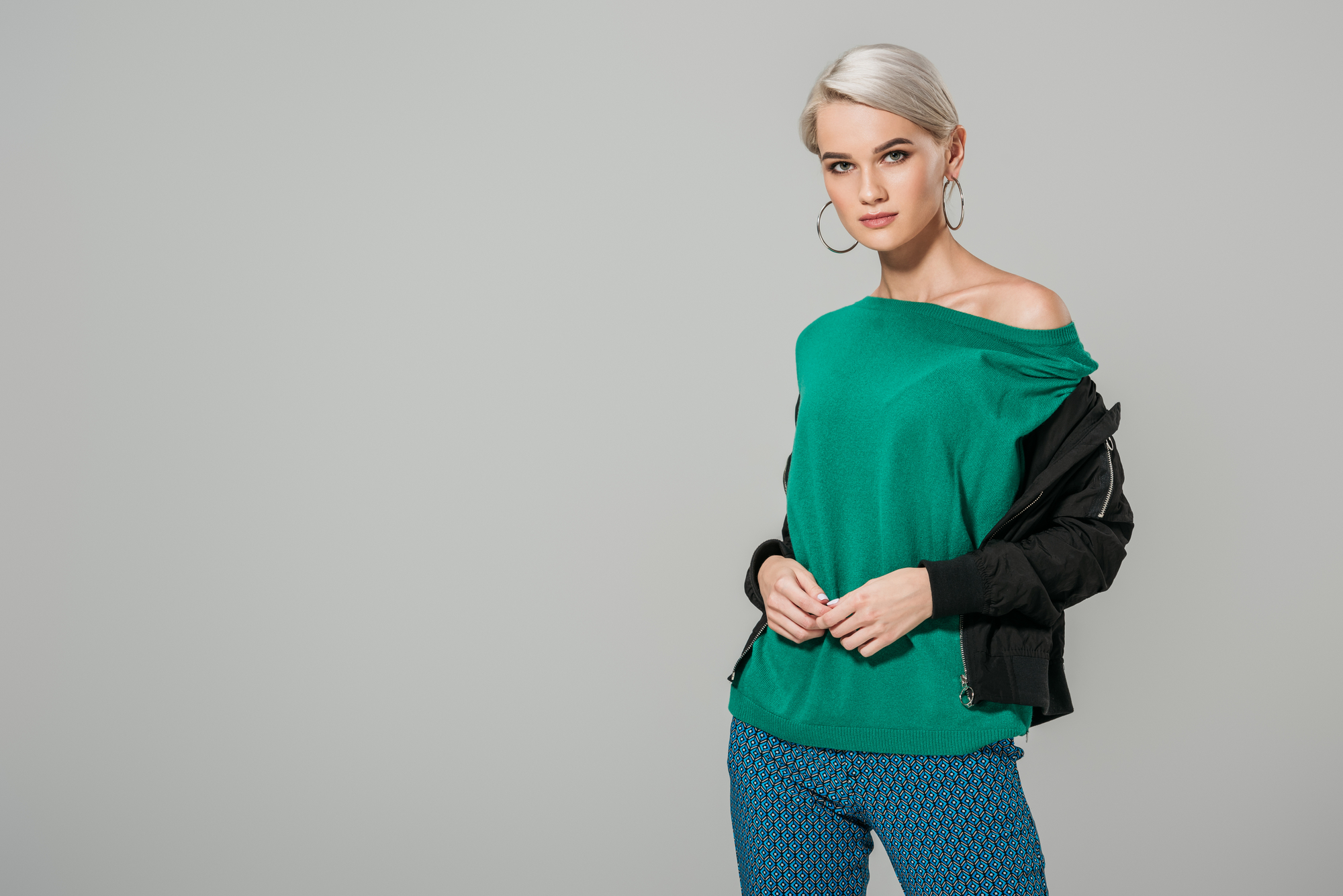 A person with short blond hair and hoop earrings stands against a plain gray background. They are wearing a green off-the-shoulder top, blue patterned pants, and a black jacket draped over their shoulders, looking confidently at the camera.