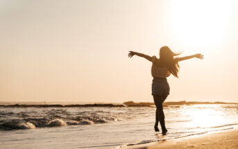 A person with long hair is joyfully running with outstretched arms along a sandy beach during sunset. The sun glows brightly in the sky, casting a warm golden light over the scene, and gentle waves are seen in the background.