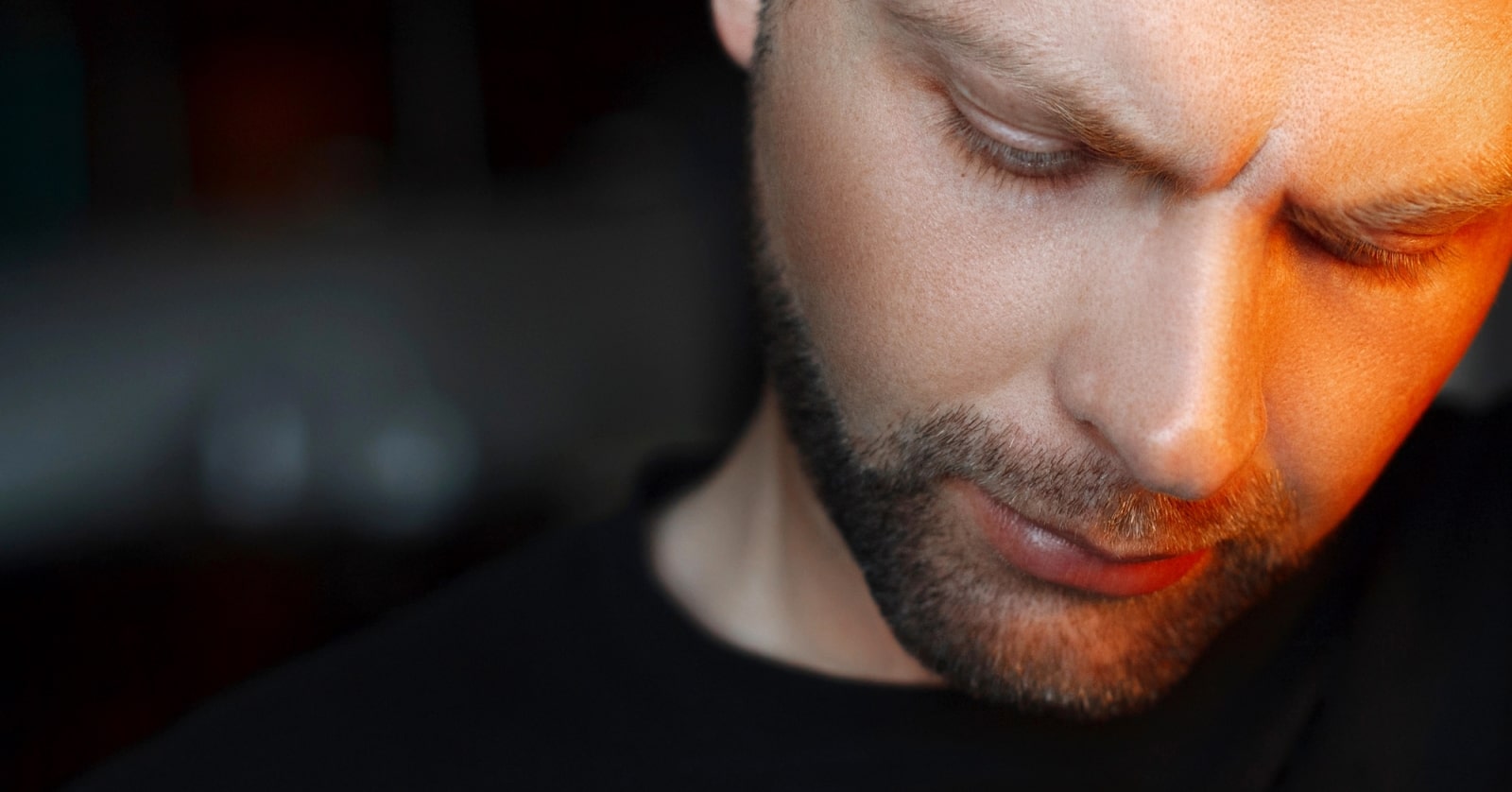Close-up of a man with a trimmed beard and short hair looking down with a serious expression. The lighting casts a warm glow on the right side of his face while the background is blurred and dark. He is wearing a black shirt.