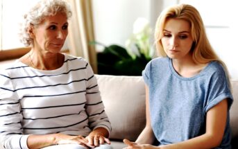 A senior woman with curly gray hair and a striped shirt sits beside a younger woman with long blonde hair and a casual blue top on a couch. Both appear to be in a serious conversation. Plants and bright light can be seen in the background.