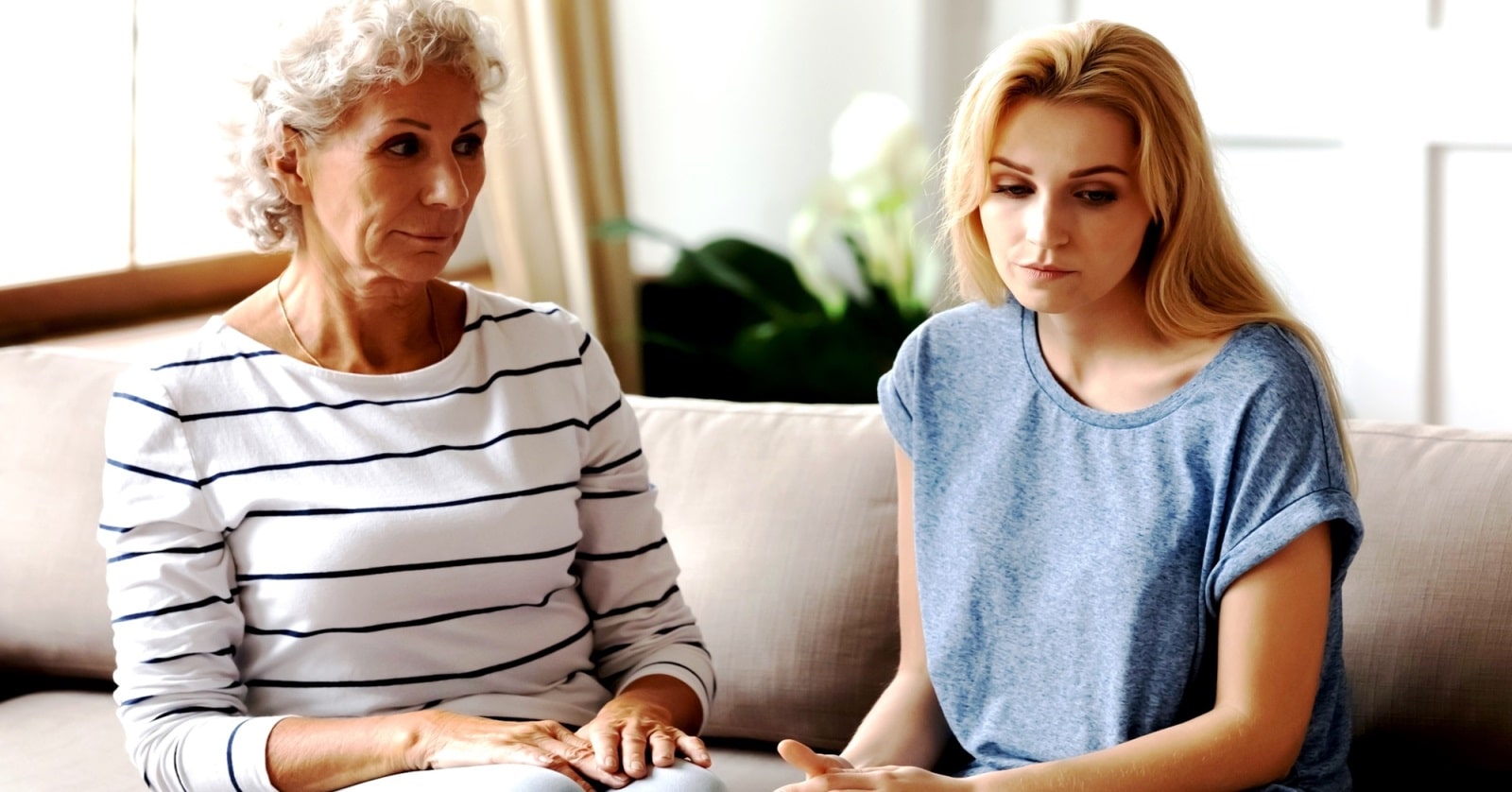 A senior woman with curly gray hair and a striped shirt sits beside a younger woman with long blonde hair and a casual blue top on a couch. Both appear to be in a serious conversation. Plants and bright light can be seen in the background.
