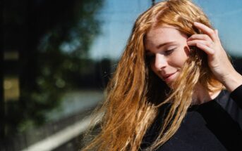 A person with long, red hair stands outdoors, lightly touching their hair with one hand. They are wearing a black long-sleeved top and appear to be in bright sunlight. The background is slightly blurred, showing trees and other natural elements.