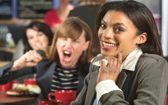 A woman in a business suit smiles and looks at the camera while sitting in a cafe. Behind her, another woman with brown hair is angrily yelling and gesturing with her fists, and a third woman watches. The scene seems to indicate conflict or aggression.