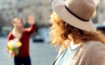 A woman with curly hair wearing a tan hat and beige cardigan stands in the foreground, facing away. In the blurred background, a man holding a bouquet of flowers waves at her. They appear to be outside, with buildings and sunlight visible in the distance.