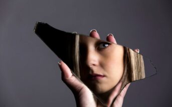 A hand holds a jagged piece of broken mirror. The mirror reveals part of a woman's face, showing her eye and partial features. The background is a plain, dark gray, allowing the reflection to stand out vividly.