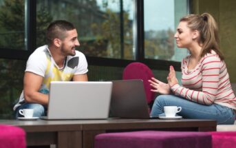 A man and a woman are sitting across a table from each other in a modern, cozy cafe. Both have laptops and coffee cups in front of them. The man is smiling, wearing a white T-shirt, while the woman is gesturing mid-conversation, dressed in a striped top.