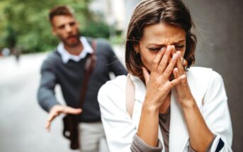 A woman with short brown hair and wearing a white coat covers her face and looks distressed, walking away along a street. Behind her, a man with a beard, wearing a grey sweater and brown shoulder bag, reaches out toward her with a concerned expression.