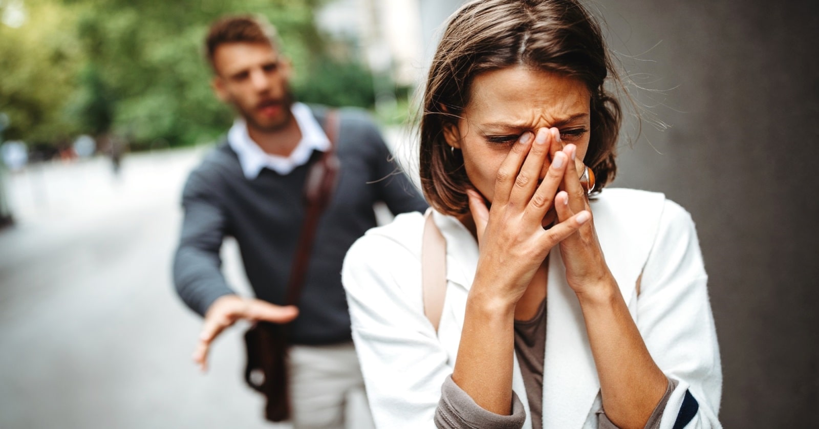A woman with short brown hair and wearing a white coat covers her face and looks distressed, walking away along a street. Behind her, a man with a beard, wearing a grey sweater and brown shoulder bag, reaches out toward her with a concerned expression.