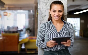 A young woman with a ponytail, dressed in a black-and-white striped shirt and green pants, stands smiling while holding a tablet. She leans against a concrete wall in a modern office space with colorful furniture and large windows in the background.
