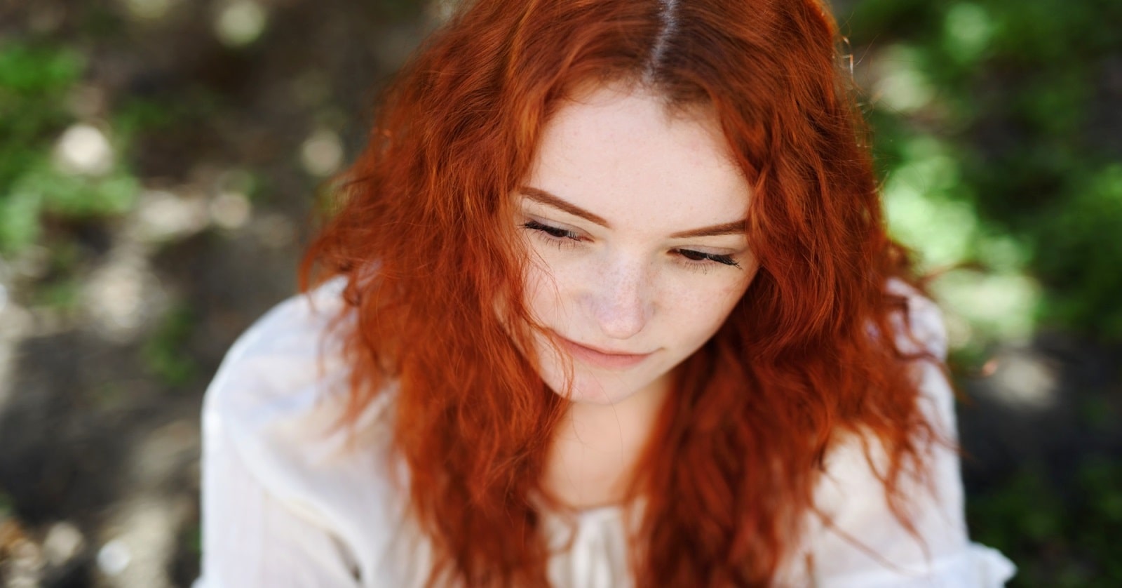 A person with long, wavy red hair and light skin looks down with a calm expression. They are wearing a white top and are outdoors, surrounded by blurred greenery. The background is softly out of focus, emphasizing the subject's serene face.