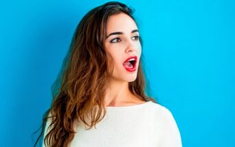 A woman with long, wavy brown hair stands against a bright blue background. She is wearing a white top and has an expression of surprise or awe, with her mouth slightly open and eyes looking off to the side.