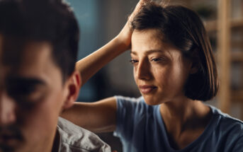 A woman with short brown hair and a sad expression places her hand on the shoulder of a man, who is facing away from her. The man appears distant, and their body language suggests tension or emotional discomfort. The setting is indoors with a blurred background.