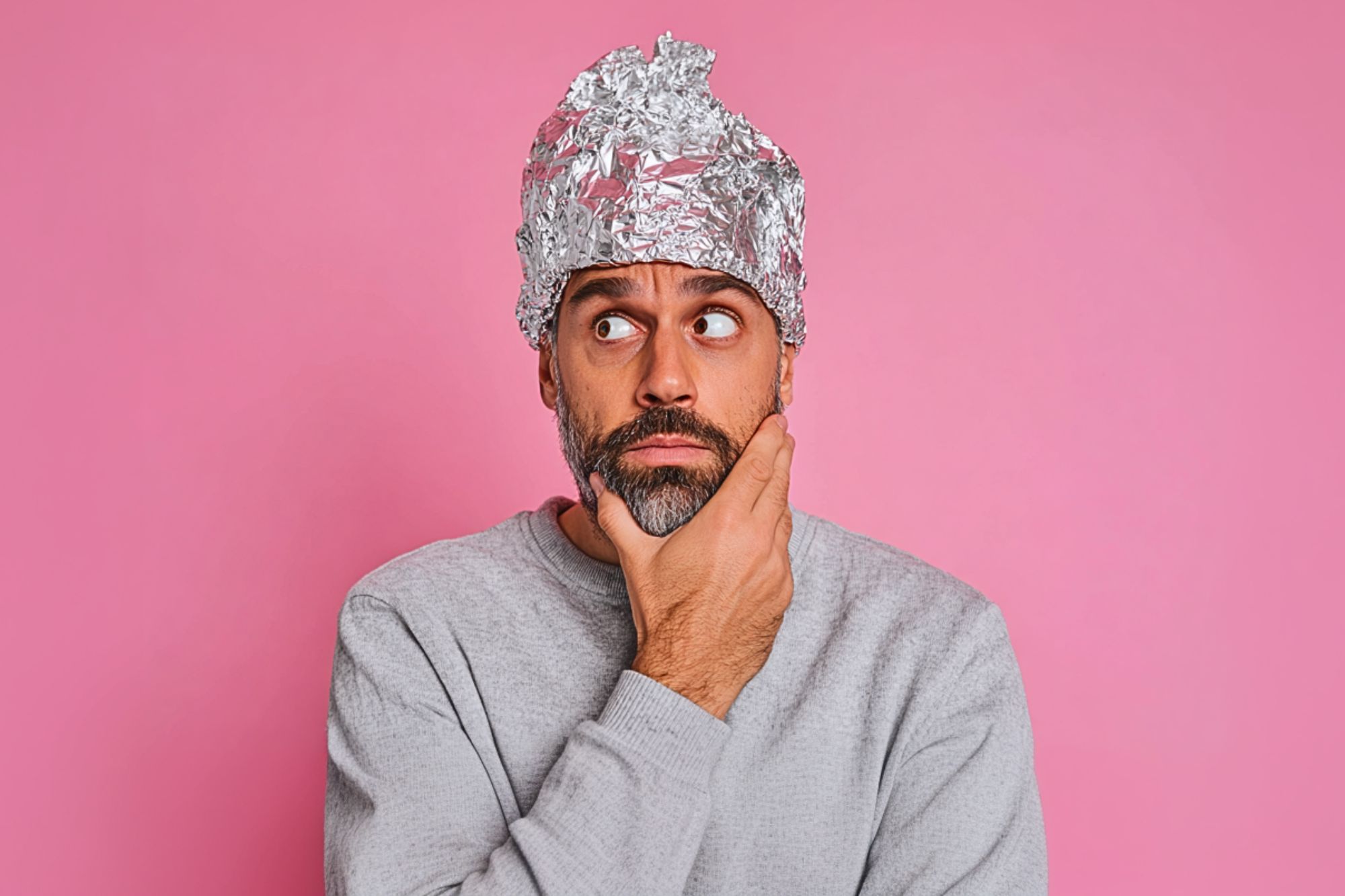 A man with a beard wearing a grey sweater and a tinfoil hat is looking to the side with a thoughtful expression, holding his chin. The background is pink.