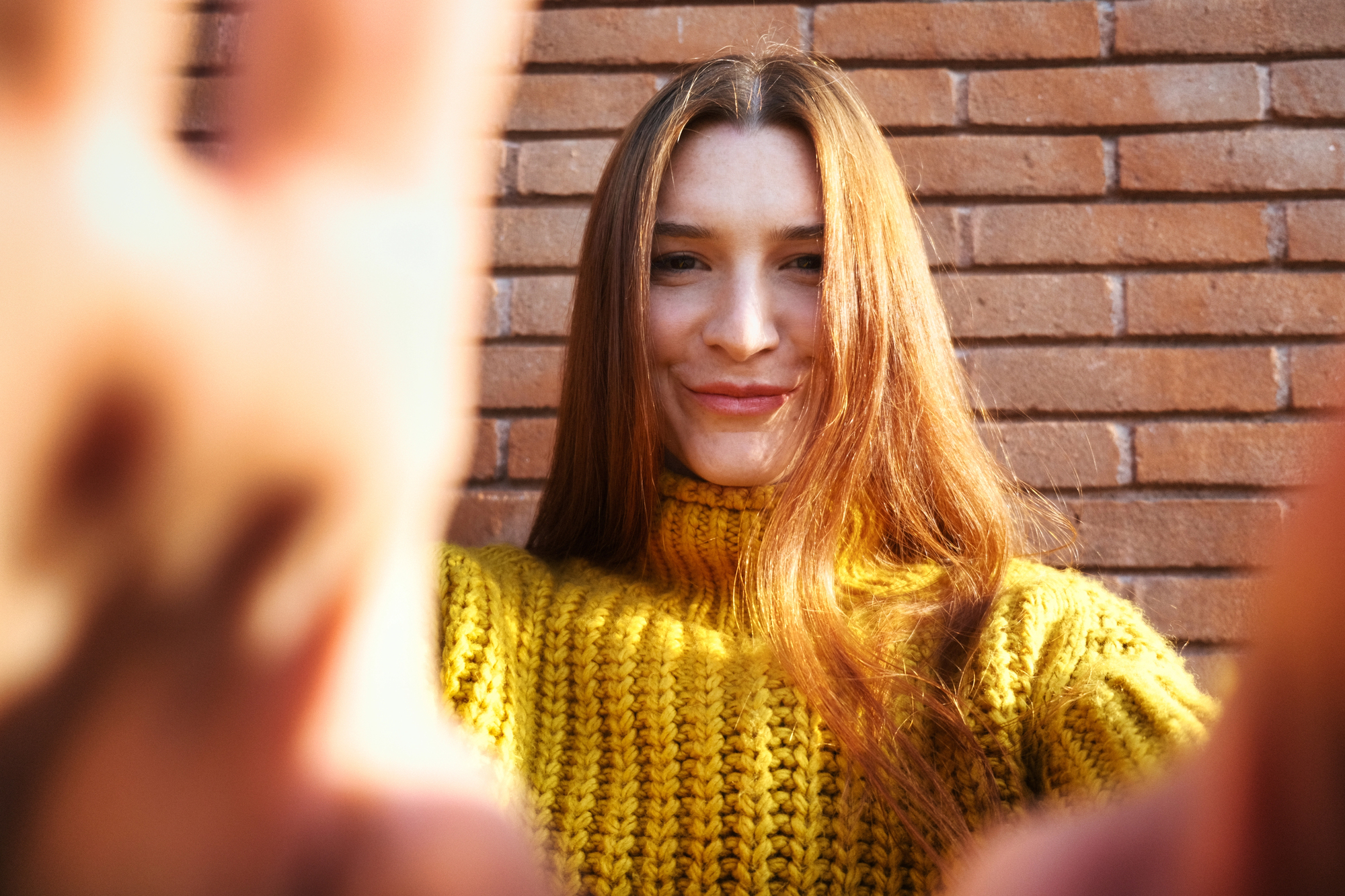 A person with long hair stands in front of a brick wall, smiling at the camera. They are wearing a bright yellow knitted sweater, and their hands are partially visible in the foreground, reaching towards the camera.