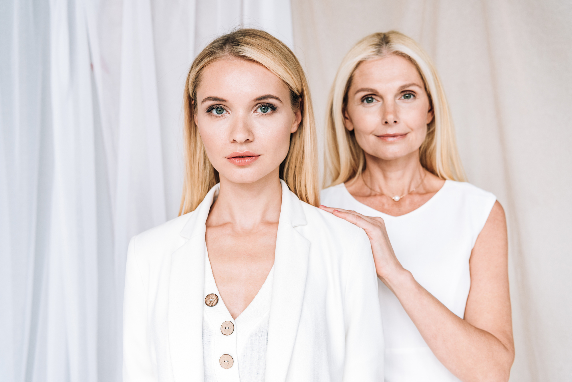 Two women with blonde hair stand together, both wearing white clothing. The younger woman in the foreground wears a blazer and looks into the camera with a neutral expression. The older woman stands behind her with one hand on her shoulder, also gazing at the camera.