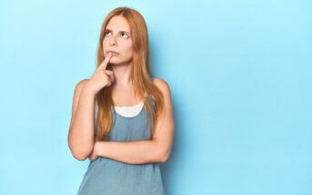 A young woman with long red hair is standing against a light blue background. She is wearing a sleeveless blue top and has one finger resting on her lips while looking upwards, appearing to be in a thoughtful or contemplative mood.
