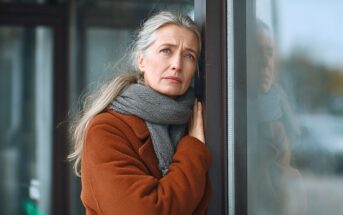 A woman with long gray hair, wearing a brown coat and gray scarf, leans against a glass window, looking thoughtfully into the distance with a serious expression.
