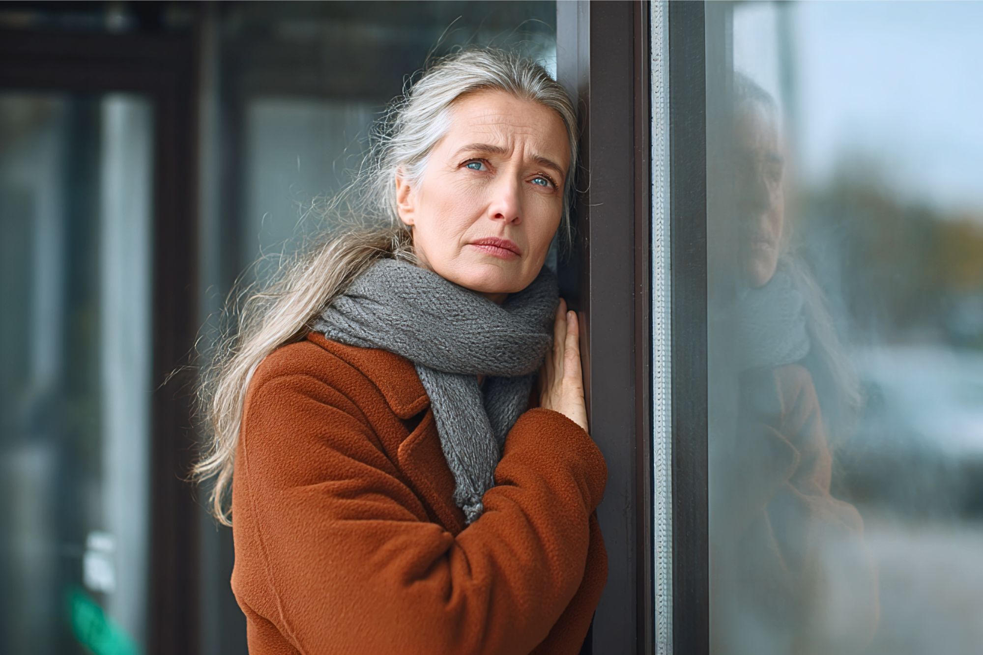 A woman with long gray hair, wearing a brown coat and gray scarf, leans against a glass window, looking thoughtfully into the distance with a serious expression.