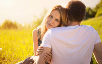 A woman with long blonde hair rests her head on a man's shoulder while sitting on a grassy field. The man, wearing a white T-shirt, faces away from the camera. Warm sunlight illuminates the scene, evoking a serene and intimate moment.