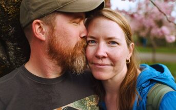 A man with a beard kisses a woman on the cheek as she smiles gently at the camera. They are outdoors, standing close together in front of blooming pink flowers.