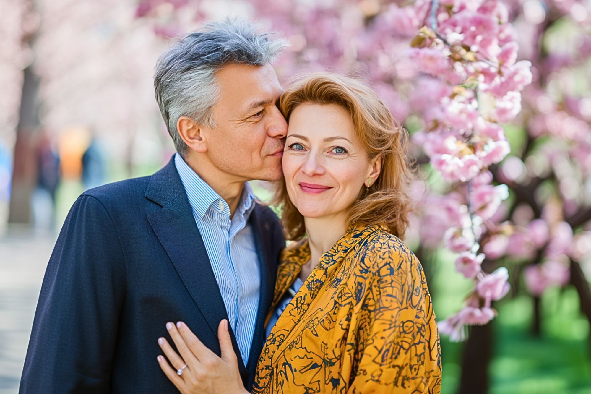 A man kisses a smiling woman on the cheek while standing outdoors among blooming pink cherry blossoms. The woman wears a patterned yellow jacket; the man wears a suit jacket and shirt.