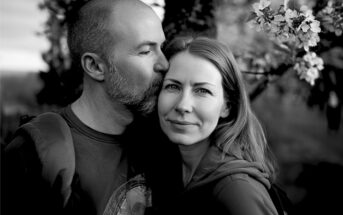 A man kisses a woman on the temple as she smiles softly at the camera. They stand close together outdoors near blossoming tree branches. The image is in black and white.