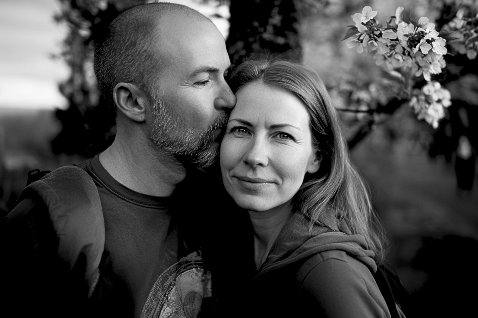 A man kisses a woman on the temple as she smiles softly at the camera. They stand close together outdoors near blossoming tree branches. The image is in black and white.