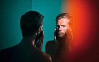 A man stands shirtless in front of a mirror, touching his face with both hands and looking at his reflection, with teal lighting and a blurred orange-red area on the right side of the image.