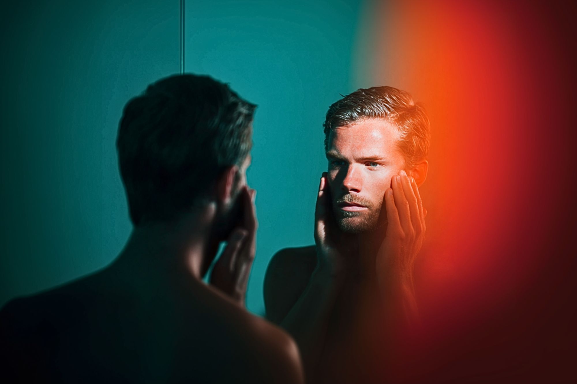 A man stands shirtless in front of a mirror, touching his face with both hands and looking at his reflection, with teal lighting and a blurred orange-red area on the right side of the image.