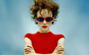 A person with short, curly brown hair wears red sunglasses and a red top, standing against a blue background with their arms crossed and an expressionless face.