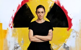 A woman in a black shirt stands with arms crossed in front of a large painted yellow and black radiation symbol on a white background.