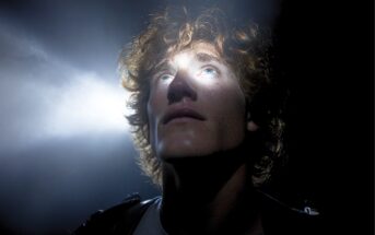 A young person with curly hair looks upward as a bright beam of light shines on their face in a dark setting, highlighting their features and creating a dramatic effect.