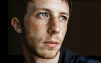 A young man with light skin, blue eyes, freckles, and short brown hair looks pensively to his left. He has a short beard and mustache. The background is dark, highlighting his thoughtful expression.