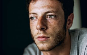 Young man with short brown hair, green eyes, and freckles gazes thoughtfully into the distance. He has light facial hair and wears a gray shirt against a dark background.