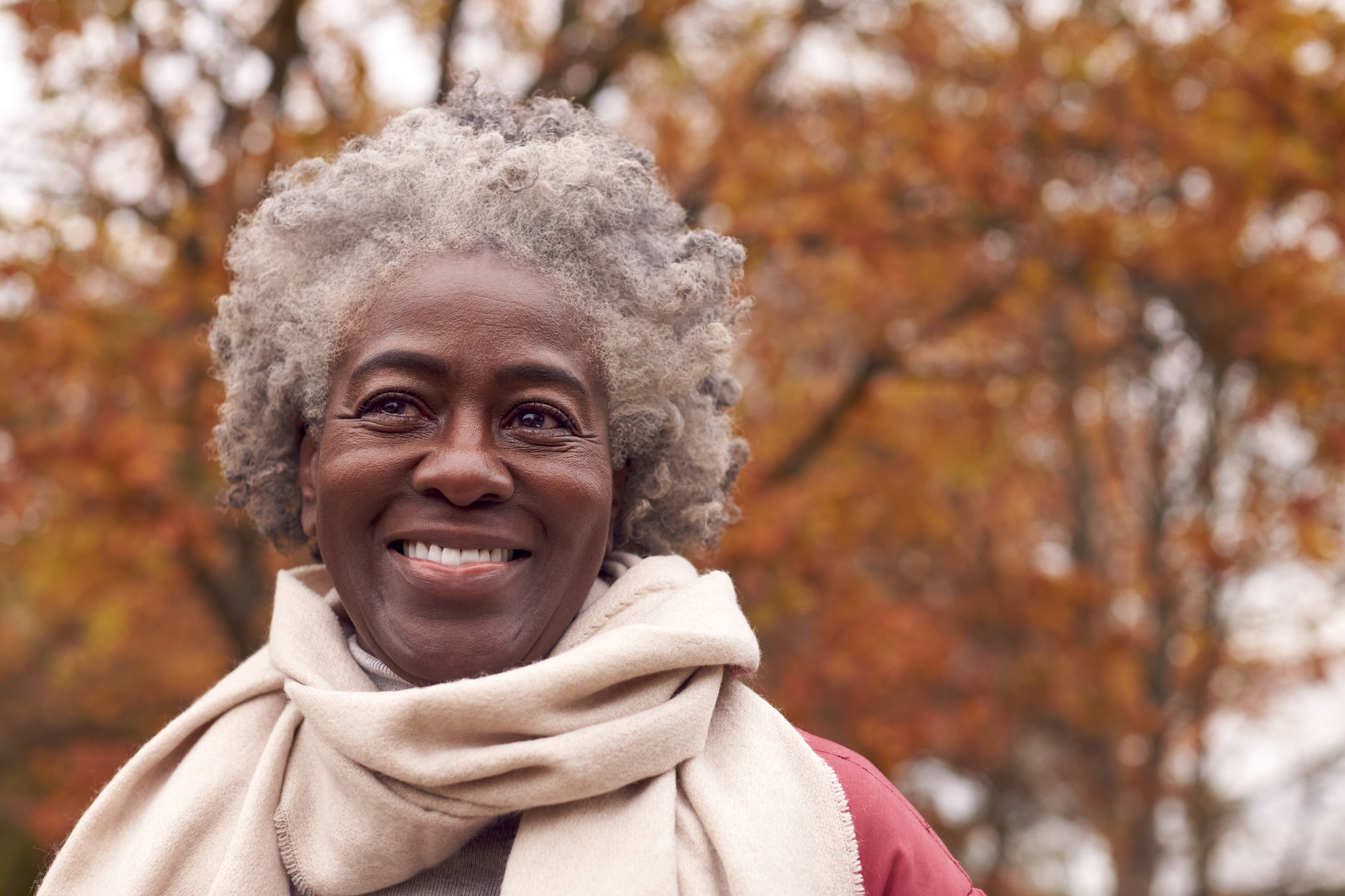 A smiling person with short curly gray hair is wearing a light-colored scarf and jacket. The background features colorful autumn trees with red, orange, and yellow leaves, creating a warm and vibrant setting.
