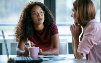 10 Disturbingly Common Behaviors That Signal Adult Bullying Two women are engaged in a conversation at an office table. One woman with curly hair and glasses is speaking, holding a pen. The other woman, with straight hair, listens attentively with her chin resting on her hand. A laptop and pink mug are on the table.