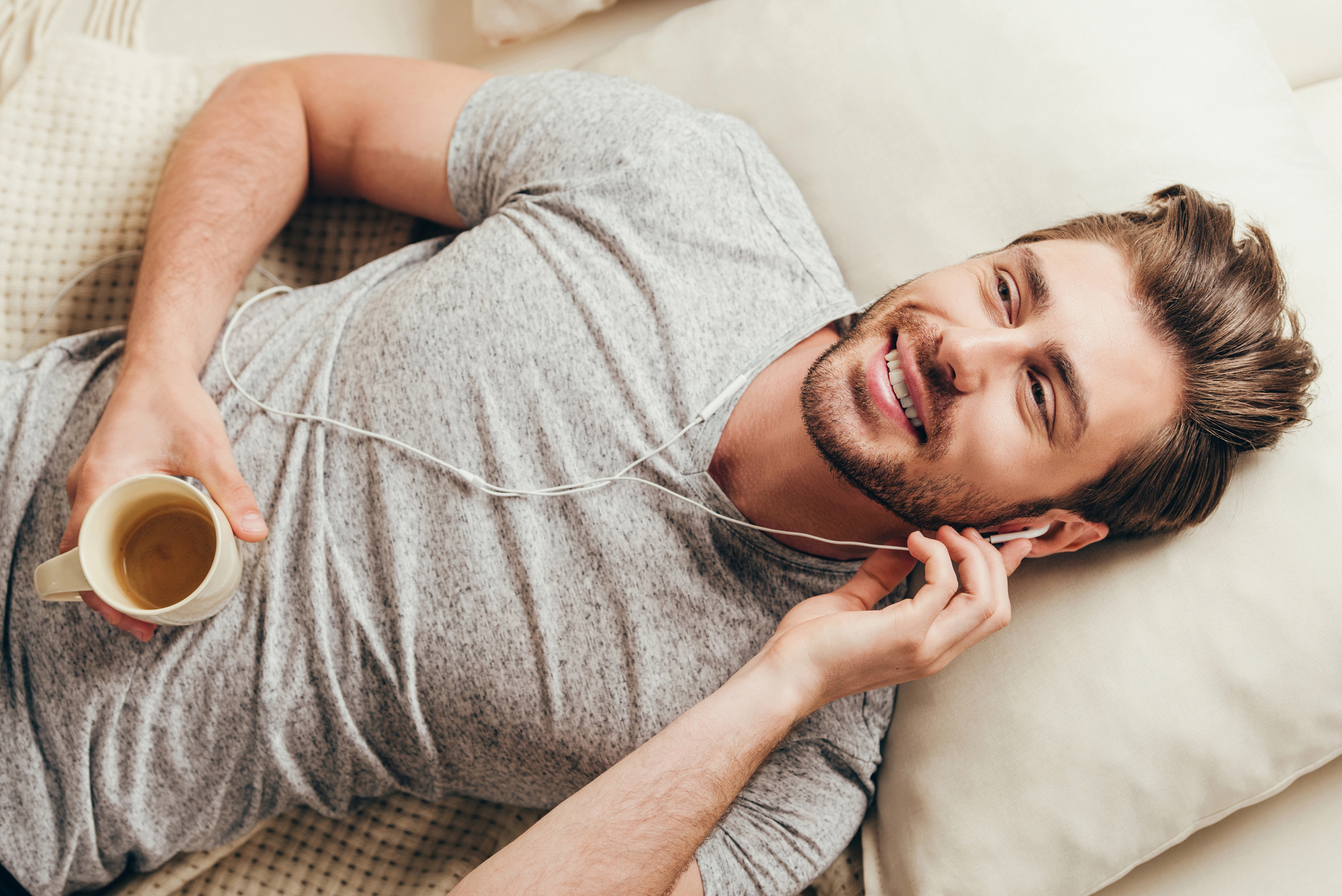A man with light brown hair and a beard is lying on a bed, wearing a gray T-shirt and white earphones. He is holding a white mug with a hot beverage in his right hand and appears to be relaxed and smiling.