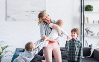 A woman smiles while holding a baby. She is standing in a bright living room. Two children, one gripping her arm and another standing nearby, interact playfully with her. The room has white walls, a gray couch, and plants and books on shelves in the background.