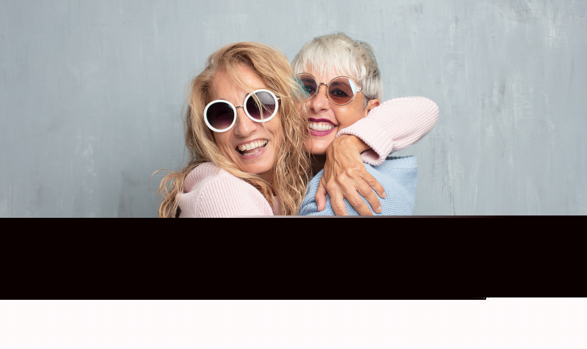 Two smiling elderly women wearing round sunglasses and cozy sweaters, embracing each other in a friendly hug against a light gray background. Both appear happy and cheerful, showing visible signs of joy and camaraderie.