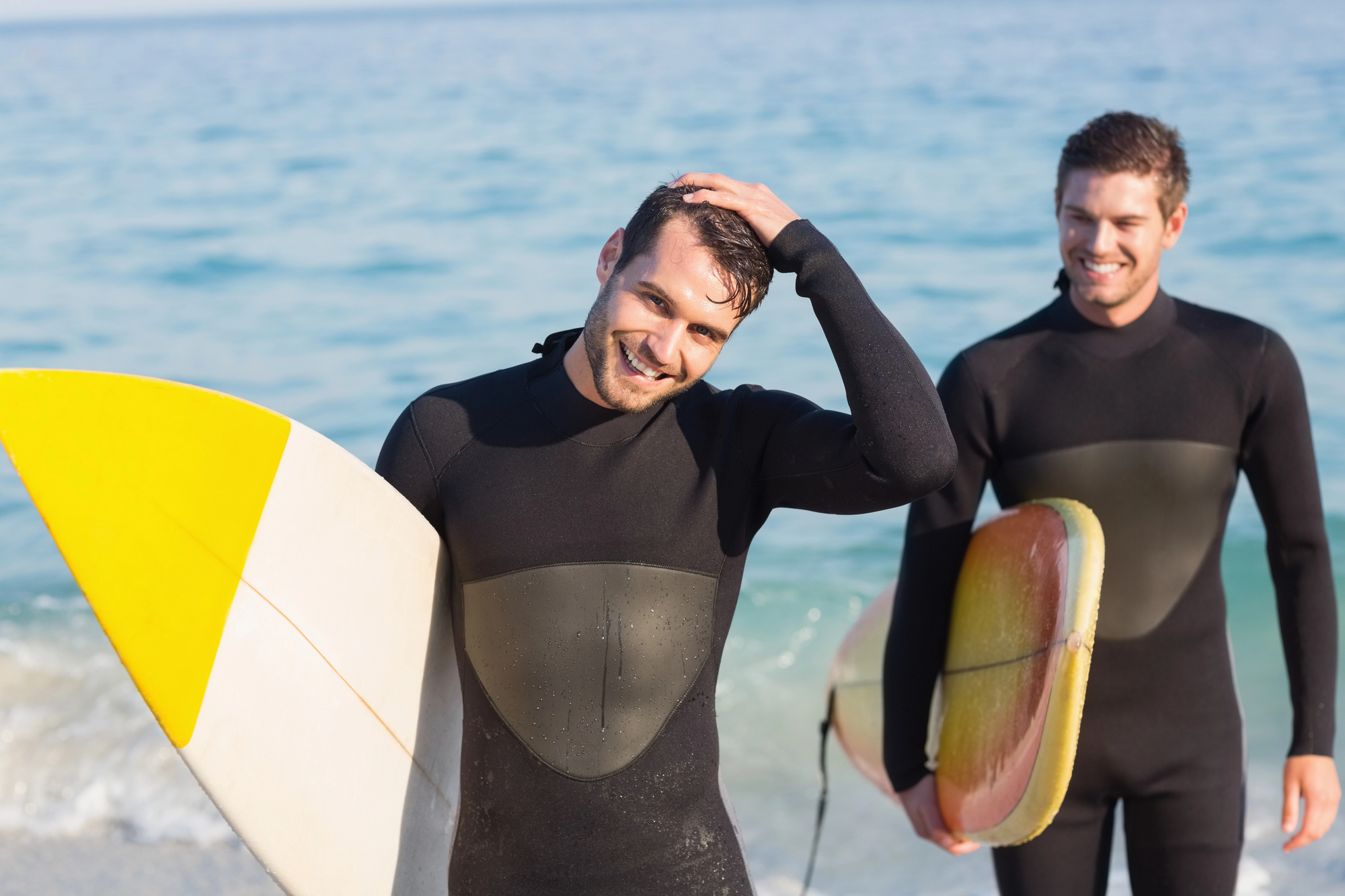 Two men wearing black wetsuits stand on a beach, each holding a surfboard. The man in the foreground has a yellow and white surfboard and is smiling while running his hand through his hair. The man behind him is also smiling, holding a red and white surfboard.