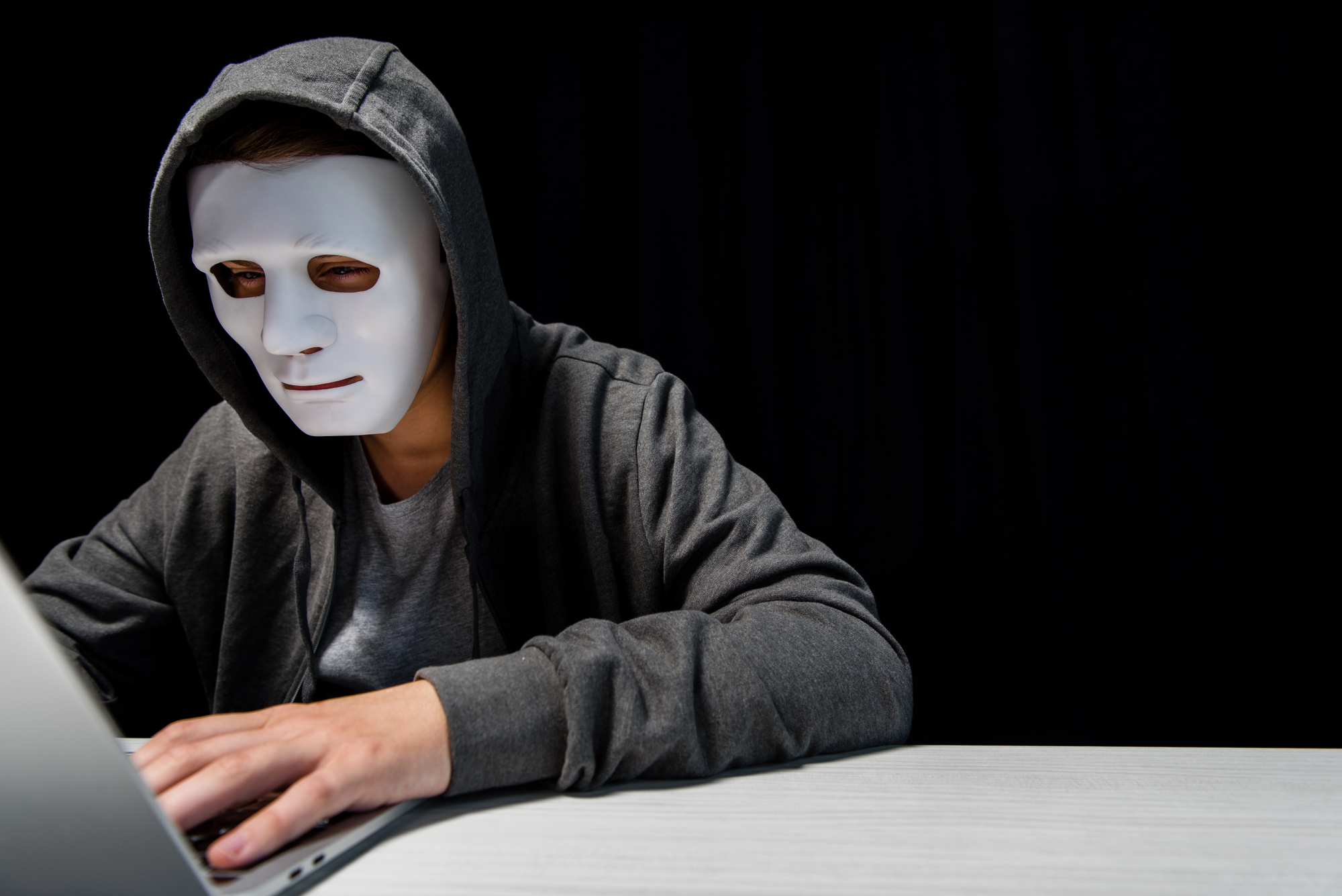 A person wearing a hoodie and a white mask sits at a table, typing on a laptop. The background is dark, adding a mysterious or possibly sinister atmosphere to the scene.